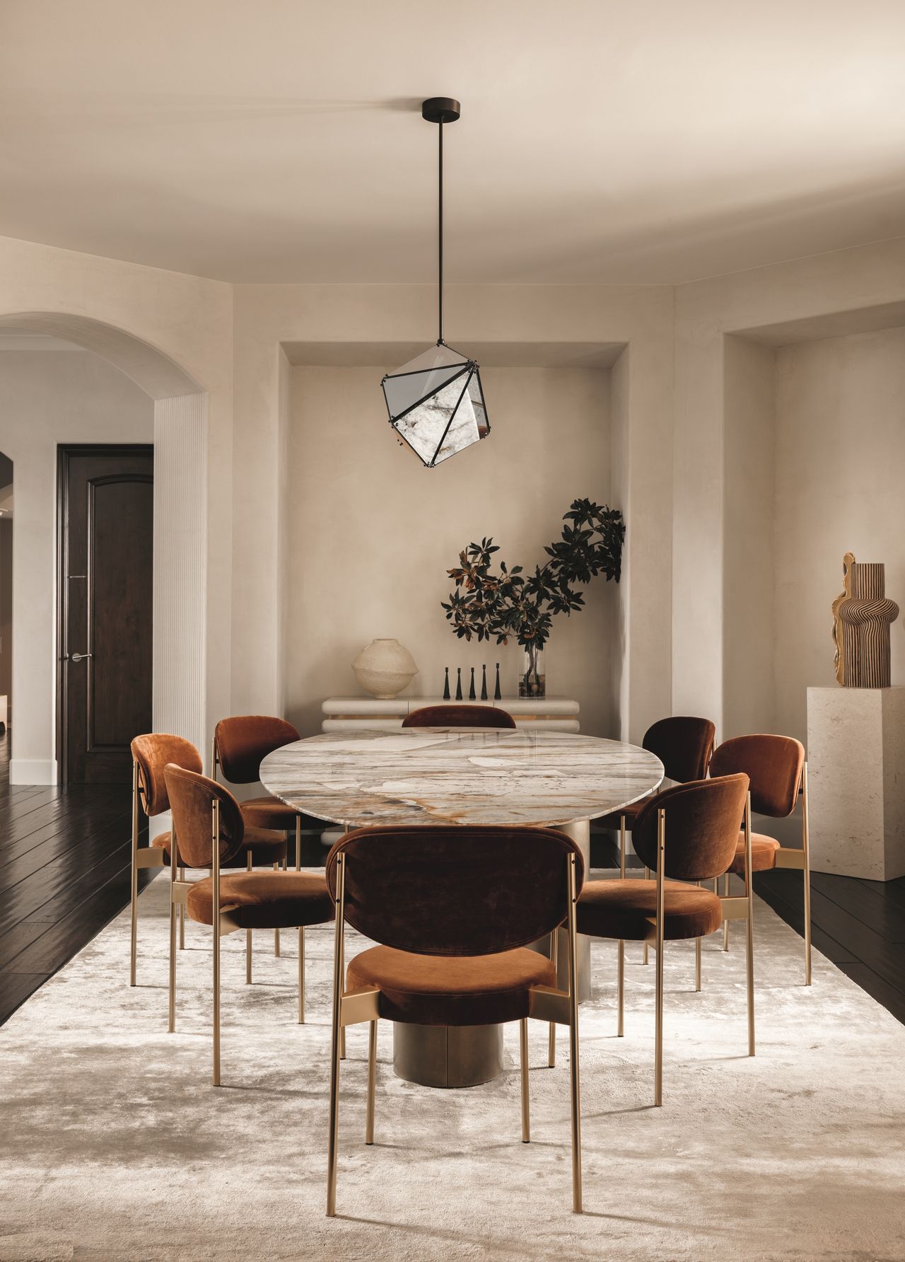 modern dining room with beige walls and ceiling with sculptural pendant light hanging over a stone-topped oval dining table with rust-velvet upholstered dining chairs and a beige velvet rug, with pedestal holding a sculpture in one corner and a console table with a sculpture and vase of foliage in the other