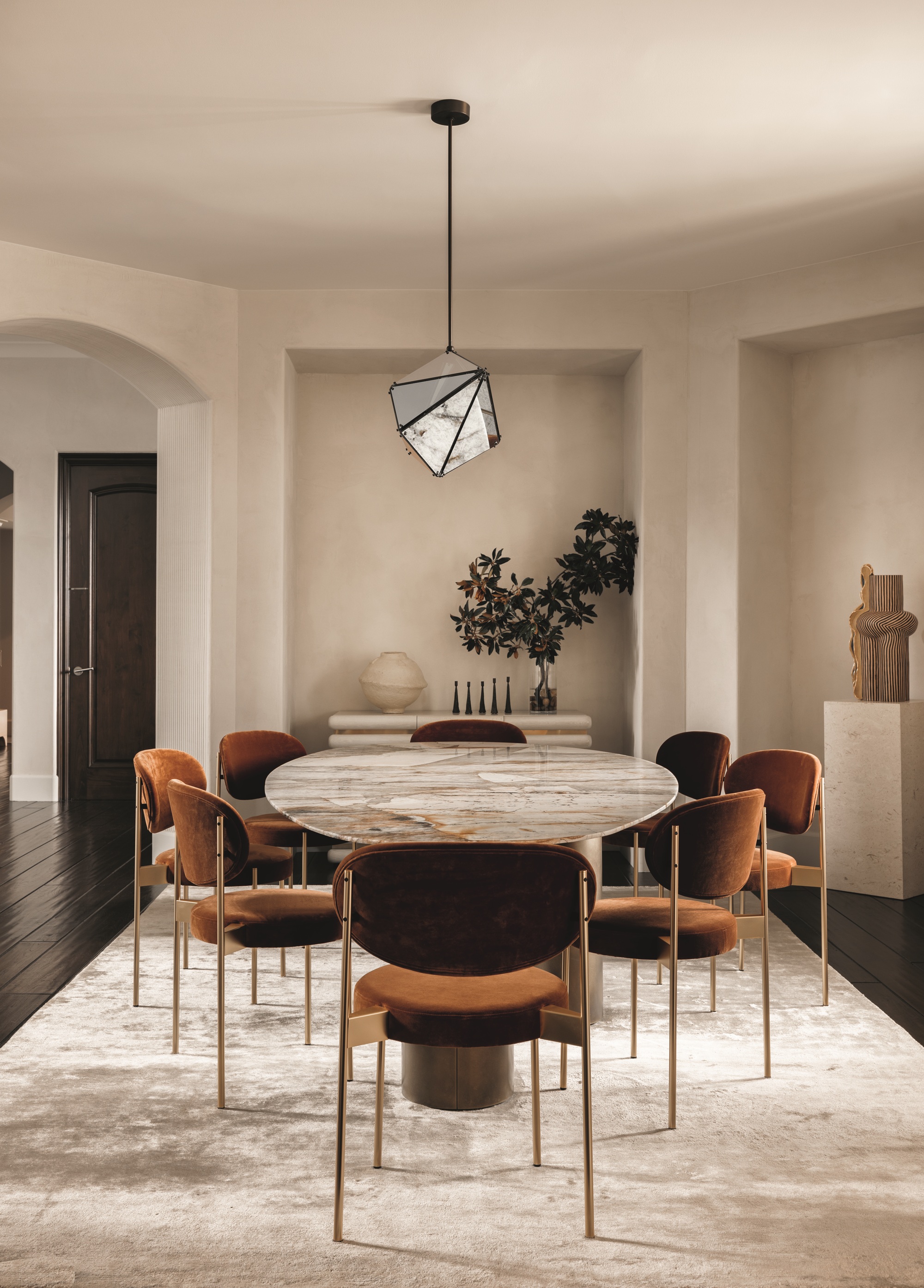 modern dining room with beige walls and ceiling with sculptural pendant light hanging over a stone-topped oval dining table with rust-velvet upholstered dining chairs and a beige velvet rug, with pedestal holding a sculpture in one corner and a console table with a sculpture and vase of foliage in the other
