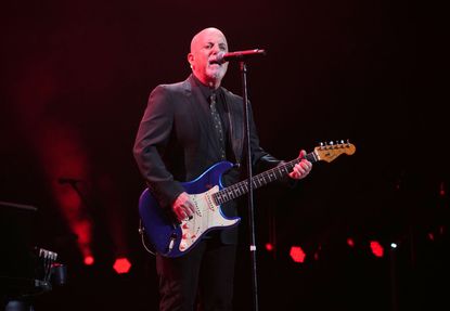 An image of singer Billy Joel wearing a suit and playing guitar on stage