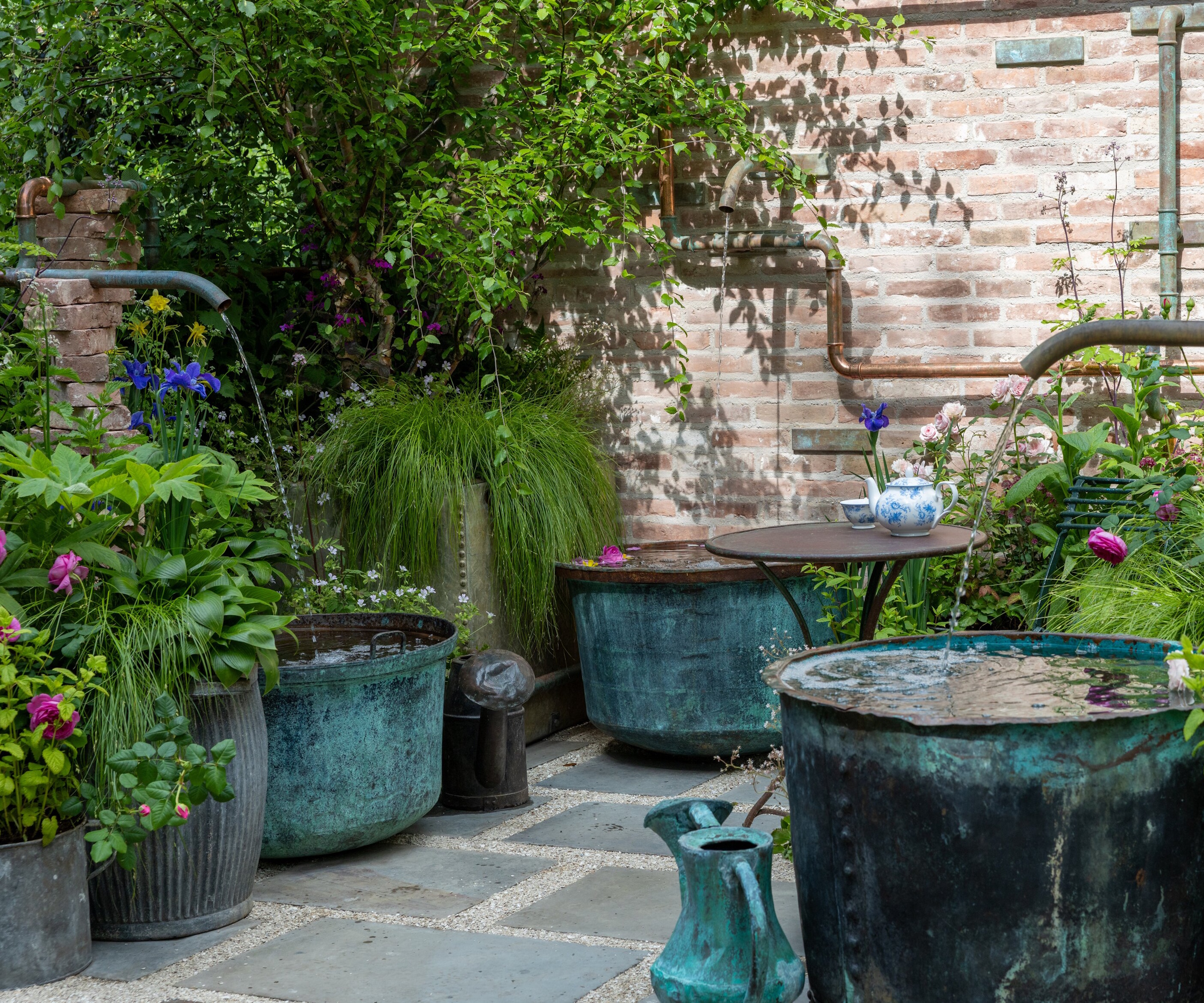 Courtyard garden with water pipes flowing into large blue copper pots and lush, cottage-style planting