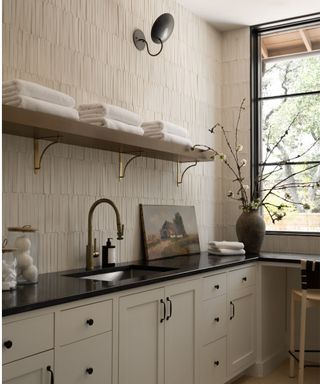 Laundry room with white tiles, cream cabinets, wooden shelf with towels and black polished surfaces with sink