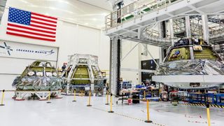 three cone-shaped spacecraft beside each other in a big white hanger. the american flag and the logo for lockheed martin are on the wall