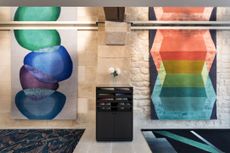 Interior view of Deirdre Dyson’s Paris showroom featuring stone walls with two colourful rugs hanging on them, stone flooring and two dark coloured patterned rugs on the floor. There is also a black unit with shelves and a cupboard with a clear vase and white flowers on top