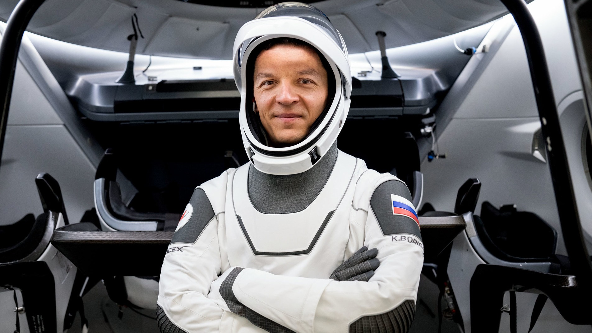 Konstantin Borisov con un traje espacial, cruzando los brazos frente a la escotilla abierta de una nave espacial