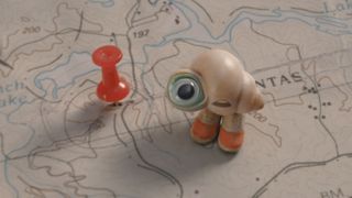 Marcel the shell with shoes on stands on a map, with a pin next to him indicating his small size