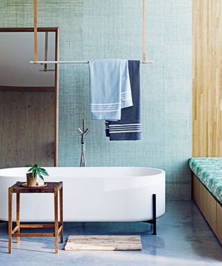 Bathroom with mint green coloured walls and screed floor, free-standing white bathtub and wooden side table, blue towels on ceiling mounted rail, window seat, wooden paneled wall