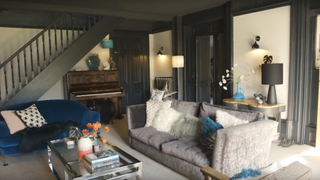 dark grey living room in a Victorian home, with a grey sofa and a blue sofa, a dark green staircase, a metal coffee table, and a piano and wooden side table against the walls