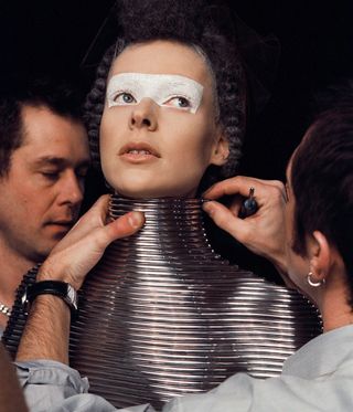 A female wearing a metallic torso top being dressed by two dressers.