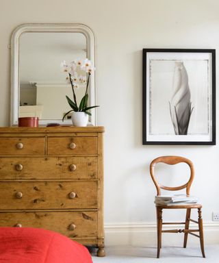 A wall of a bedroom with a curved white mirror above a rectangular set of drawers with an orchid on top, a black and white wall art print above a wooden curved chair with gray cushioning, and the corner of a red bed