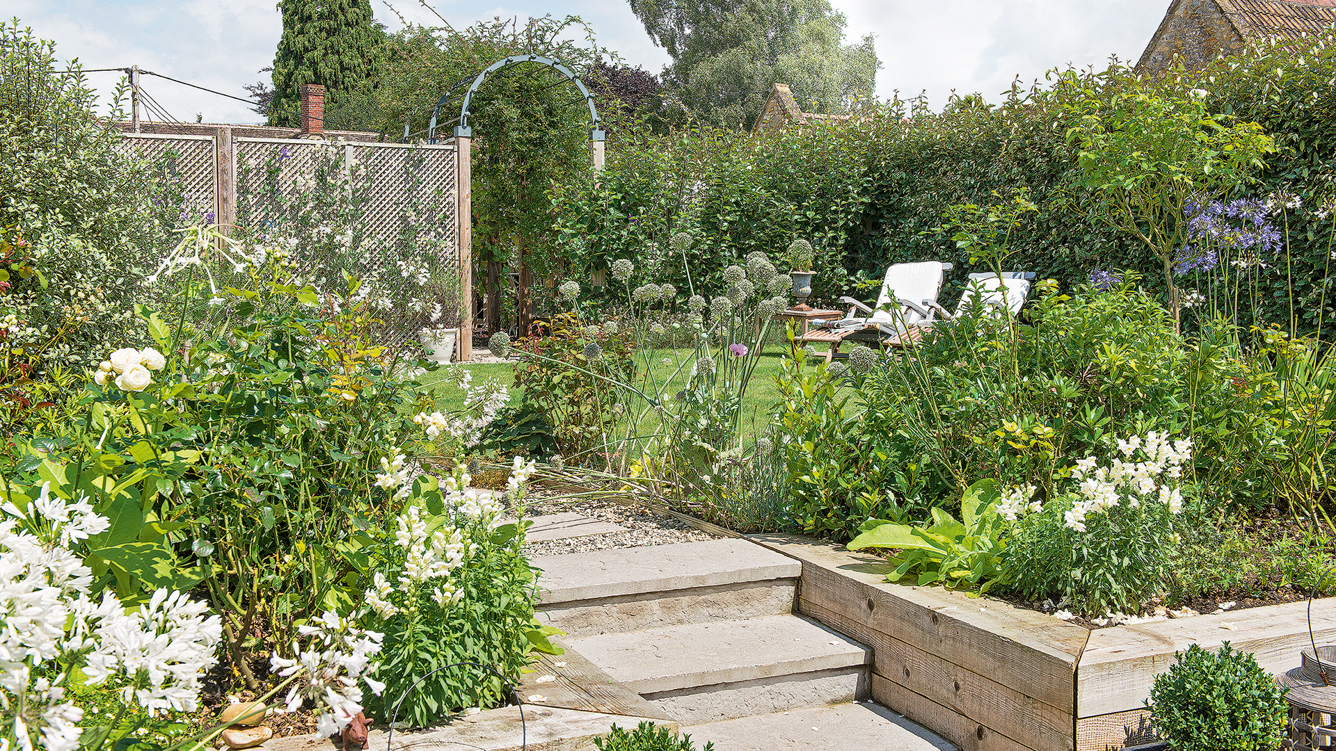 A split level garden with raised flower beds with white flowers surrounding a lawn with two white sun loungers
