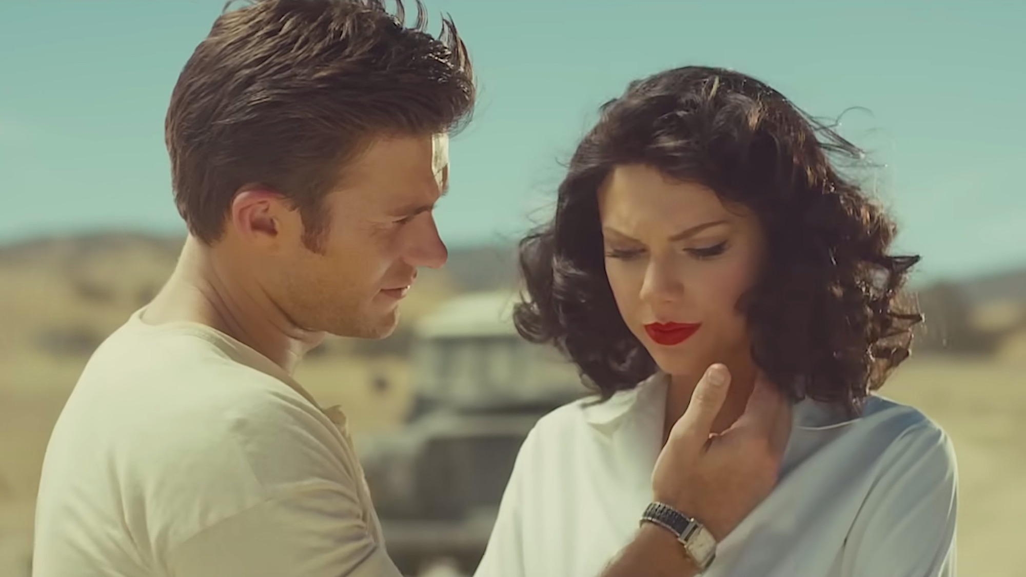 Scott Eastwood reaching out to touch Taylor Swift's face in Wildest Dreams music video. Swift is wearing a brown wig and they are in the desert.