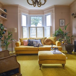 carpet colour trends for 2023, living room with blush walls, yellow carpet, footstool and sofa, rattan chair, plants, cushions