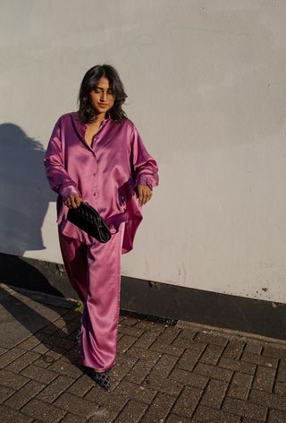 Hanukkah outfit idea is shown in a photo of a woman standing outside wearing a pink satin pajama set with crystal mesh gucci heels and a black clutch bag