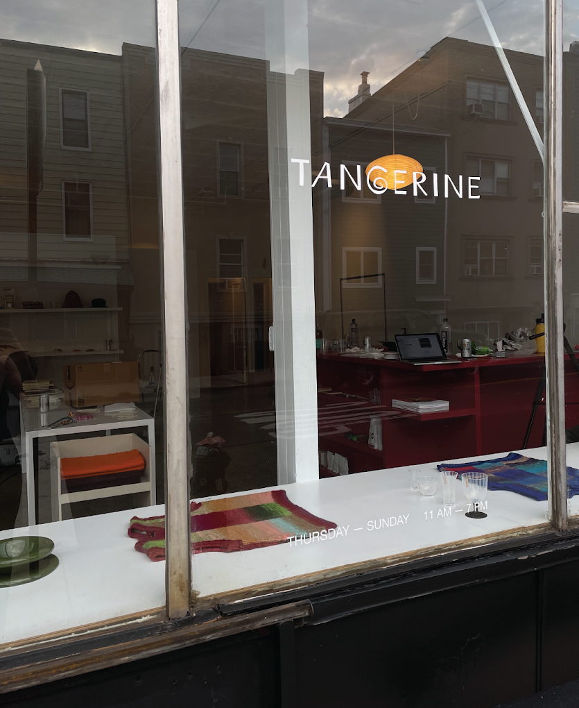 An independent store's front features an handwritten-like white logo that reads "TANGERINE". Inside, a series of colorful garments and furniture pieces.