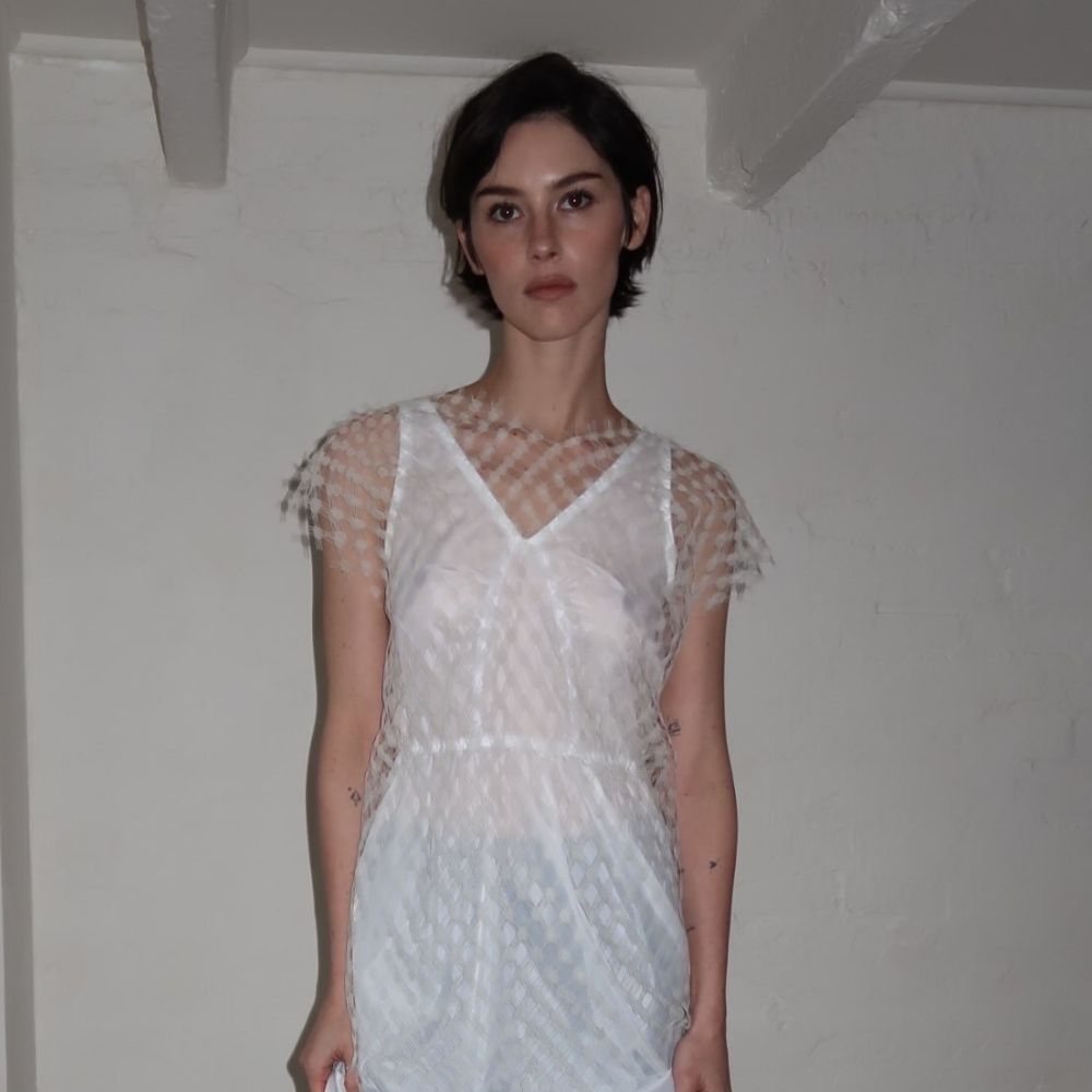 Gracie Abrams poses for a photo wearing a white semi-sheer minidress with opaque black tights and white pointed-toe heels.
