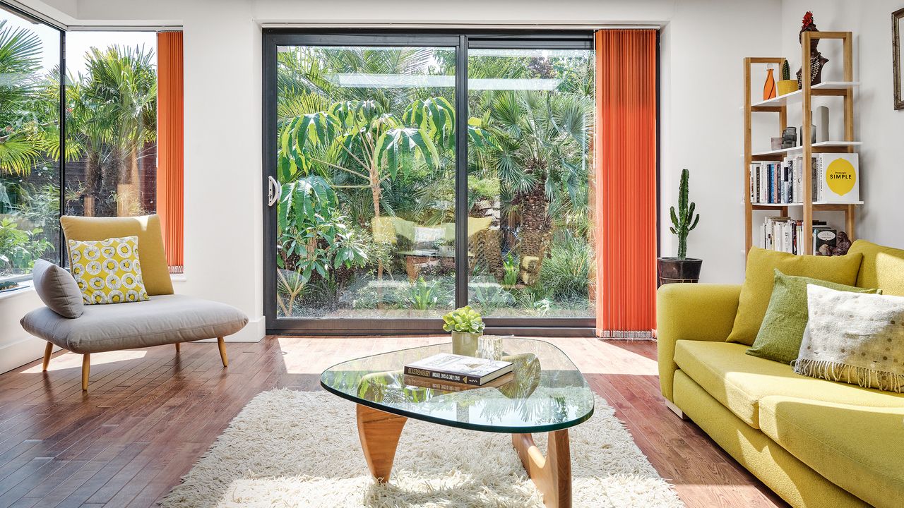 White living room with yellow sofas, warm wood flooring and sliding doors looking out onto an exotic garden