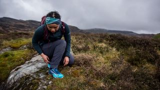 A woman on a mossy hillside crouches down to look at her Dynafit Feline SL Running Shoes.