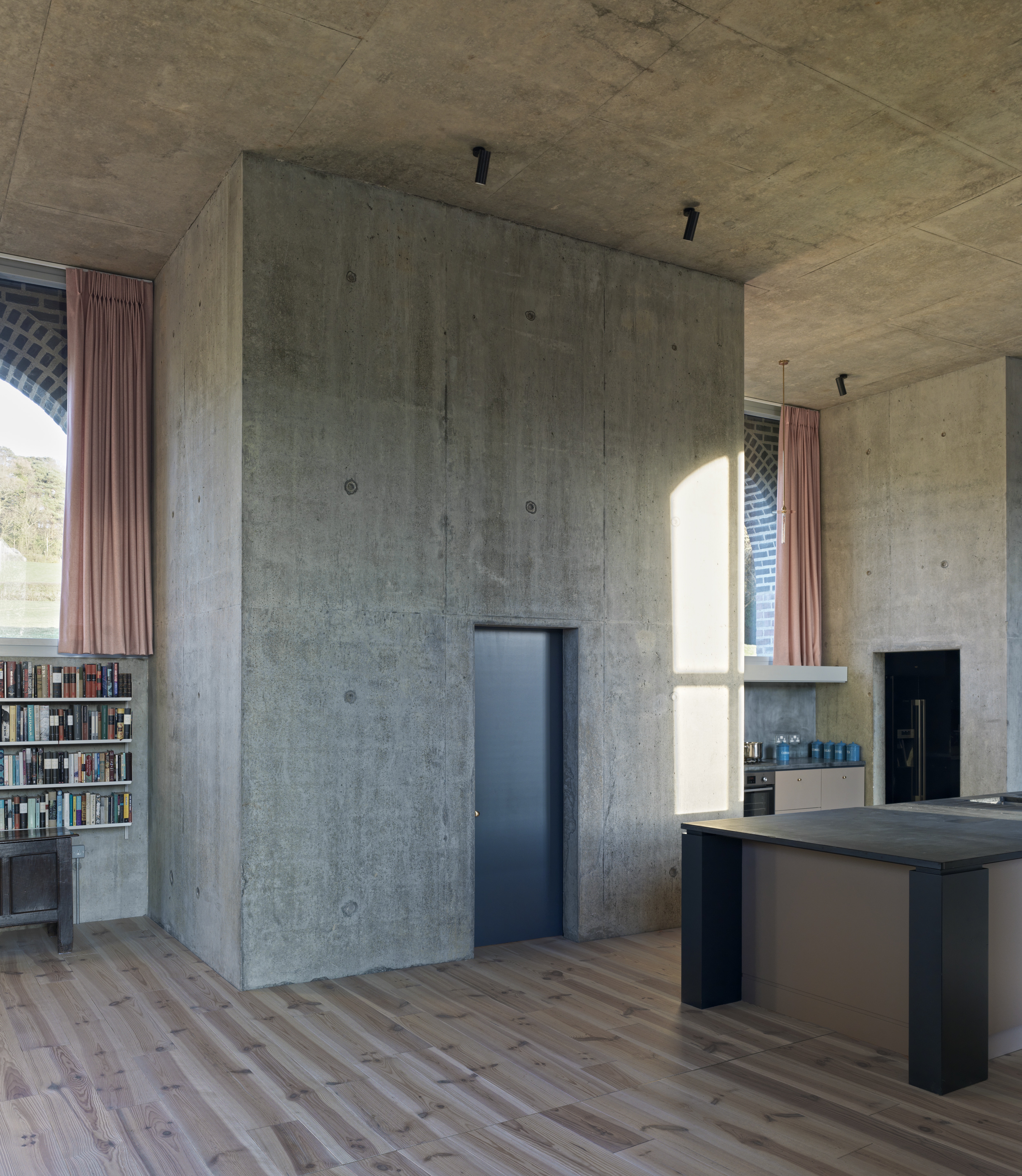 A kitchen area with a black island counter, a stove, an oven, storage cupboards, a bookshelf, a high roof and large windows.