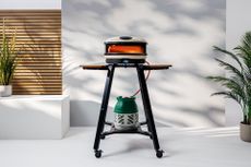 Gozney's Arc XL pizza oven lit up against a white background with plants either side and the sun casting shadows against the back wall