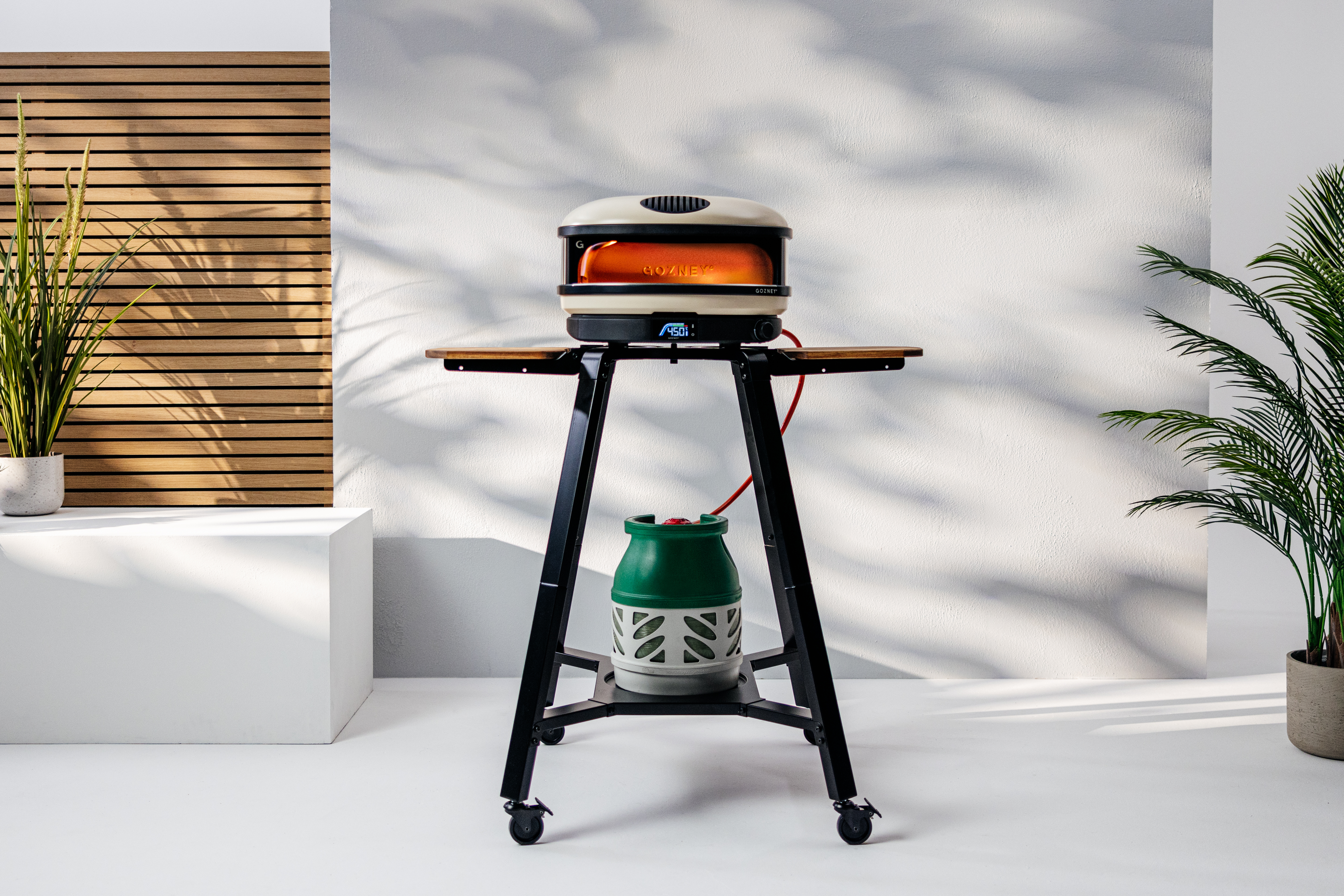 Gozney&#039;s Arc XL pizza oven lit up against a white background with plants either side and the sun casting shadows against the back wall