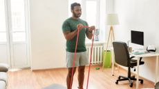 Man in domestic setting performs biceps curl with a long looped resistance band. In one corner of the room is a couch, a desk in the opposite corner. The man wears grey shorts and a green T-shirt. He has short black hair and a beard