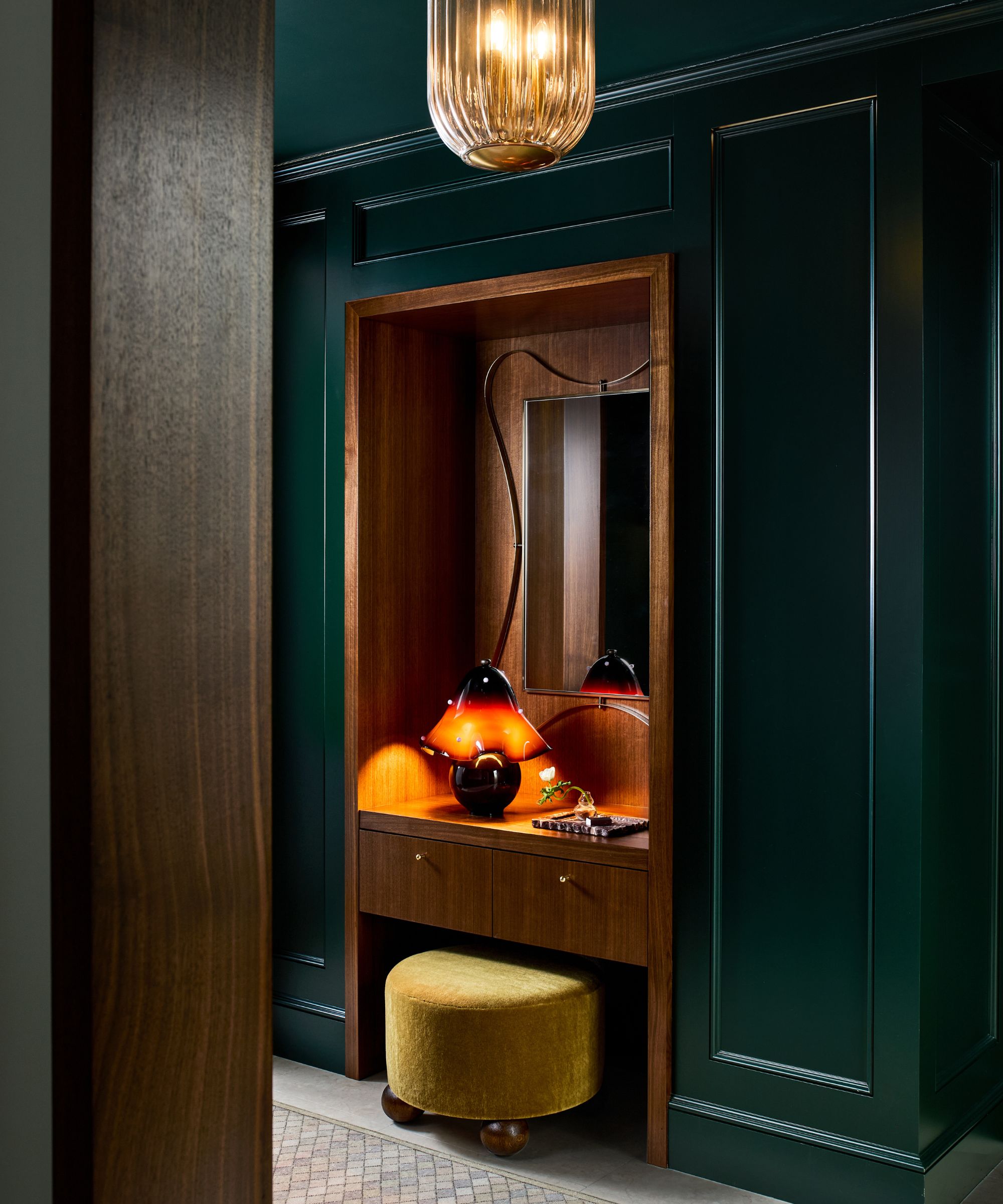 A luxurious built-in wood vanity set into a deep emerald green paneled wall, featuring a glowing amber mushroom lamp and a chartreuse velvet ottoman