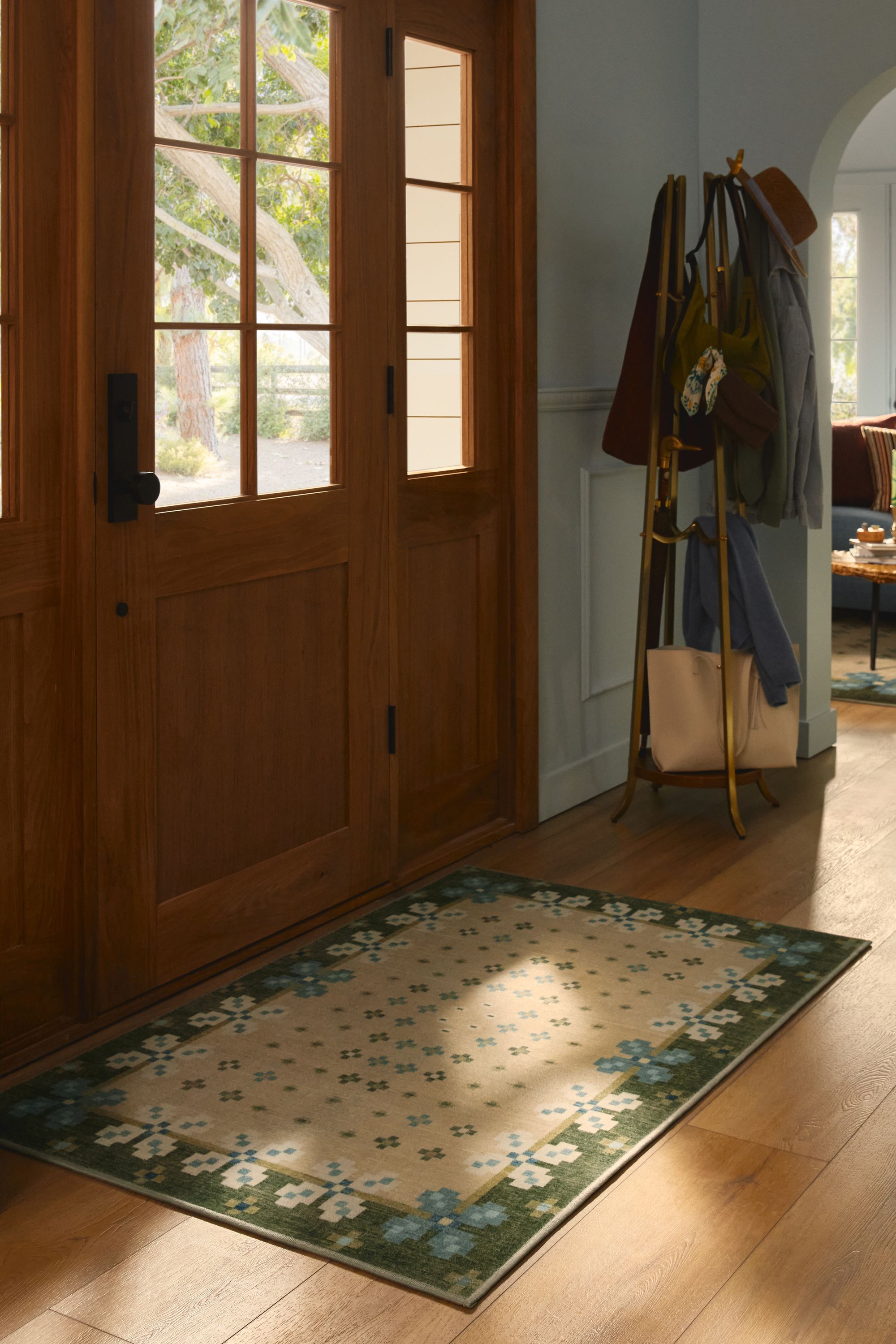 Image of an entryway with a wooden door, wooden floors, light blue walls, a coat stand, and a green Scandinavian printed rug.
