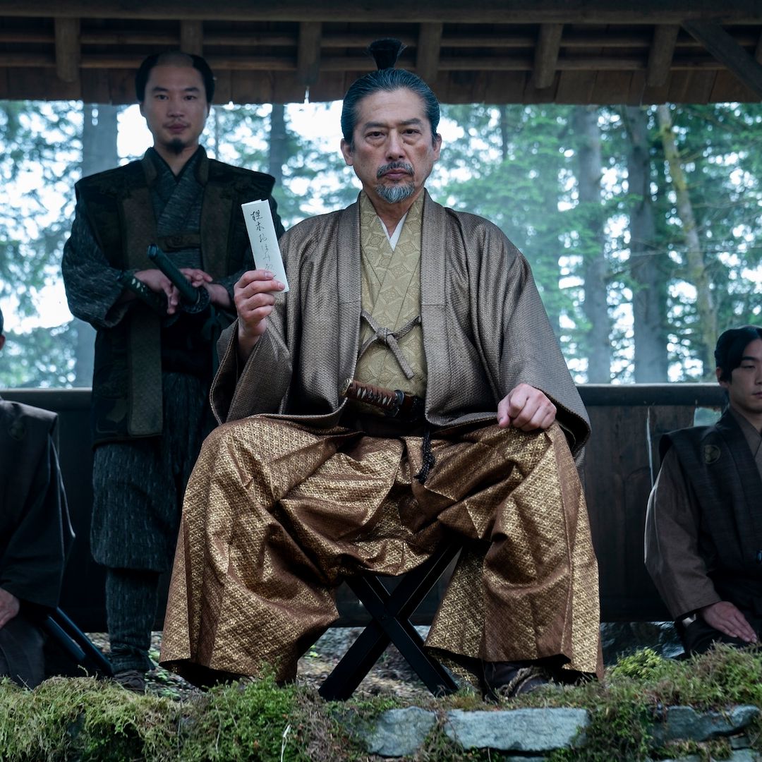 a man in gold japanese period wear (Hiroyuki Sanada as Yoshii Toranaga) sits while holding up a white letter, with one man standing behind him and two more kneeling at each side, in &#039;shogun&#039; (2024)