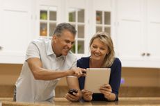 A happy, middle-agedcouple using digital tablet and cell phone at kitchen counter.