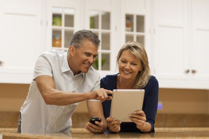 A happy, middle-agedcouple using digital tablet and cell phone at kitchen counter.