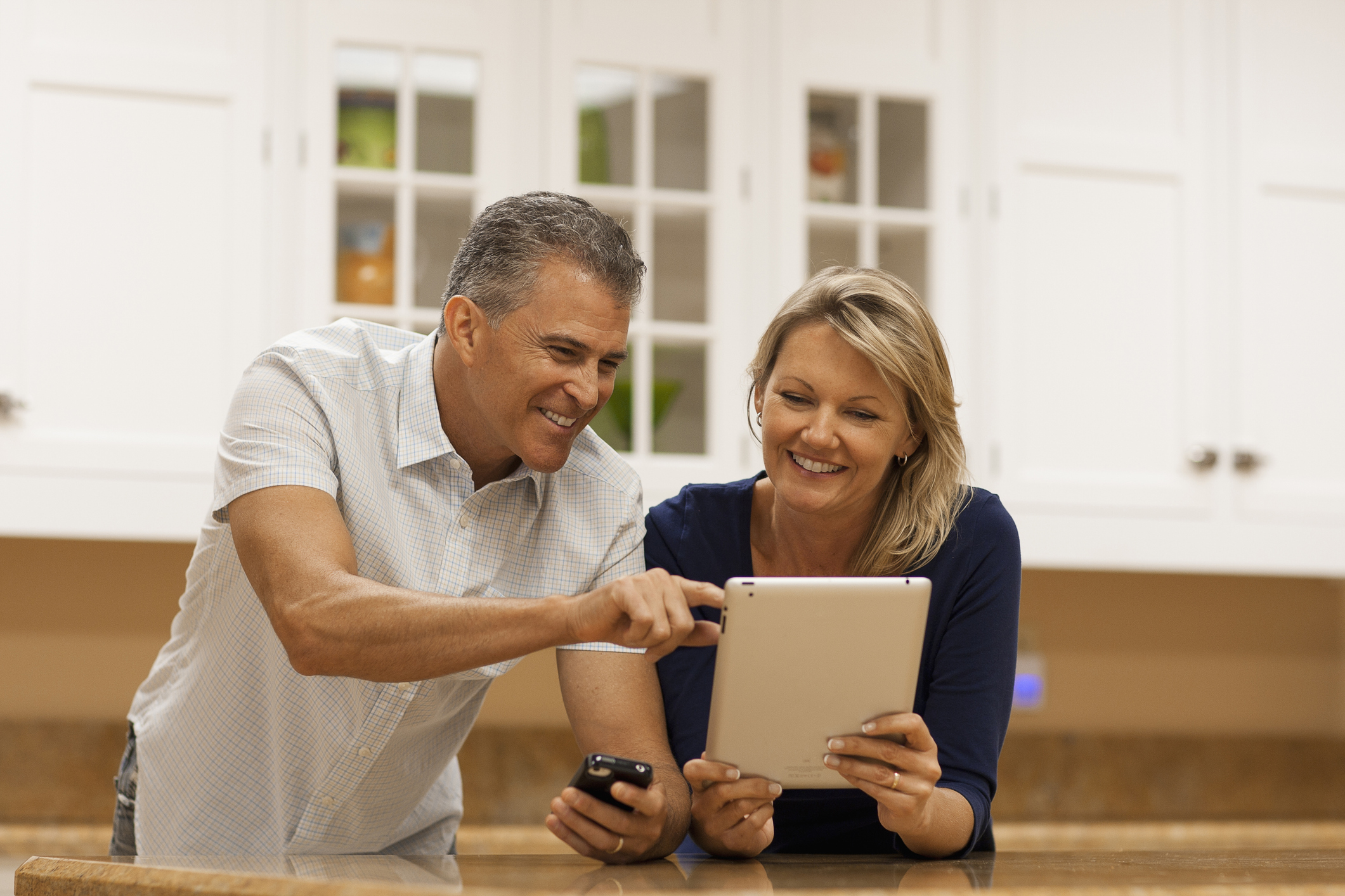 A happy, middle-agedcouple using digital tablet and cell phone at kitchen counter.