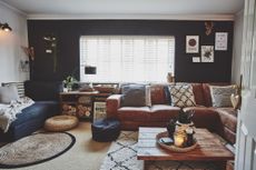 Living room with one dark black/blue wall, other white walls, tan leather L-shape sofa, black fabric sofa, rugs, rattan pouffs and coffee table made from wooden palett boards perfect for a cosy night in