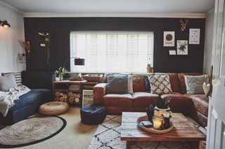 Living room with one dark black/blue wall, other white walls, tan leather L-shape sofa, black fabric sofa, rugs, rattan pouffs and coffee table made from wooden palett boards