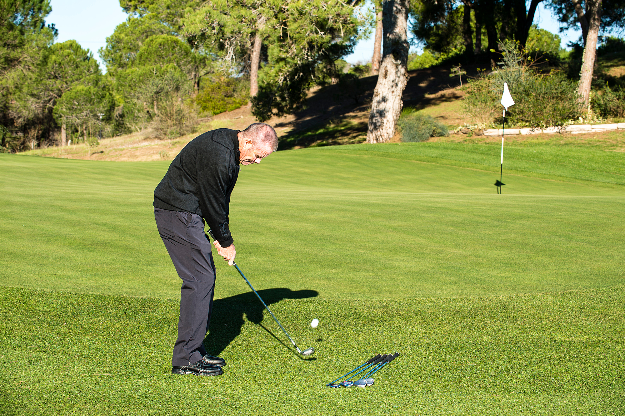Gary Alliss chipping from off the green with a wedge, with the ball coming off the clubface and travelling towards the hole, with three other clubs laid on the ground next to him as alternate options for club choice