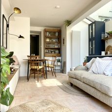 white living room with pale terracotta floor tiles &ndash; a small dining table and wooden chairs in the background and a cream sofa in the foreground 
