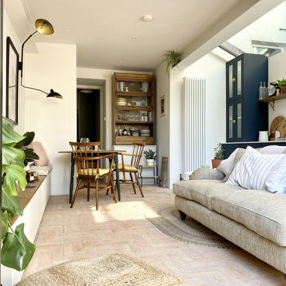 white living room with pale terracotta floor tiles – a small dining table and wooden chairs in the background and a cream sofa in the foreground 