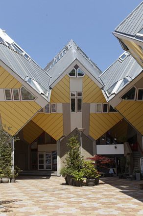 Close up exterior view of Rotterdam's Cube Houses during the day. The houses are yellow and grey cubes that are tilted 45 degrees. There is a patterned multicoloured floor outside and green plants in pots