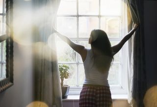 A woman pulls back the curtains in the morning to allow natural sunight into her bedroom