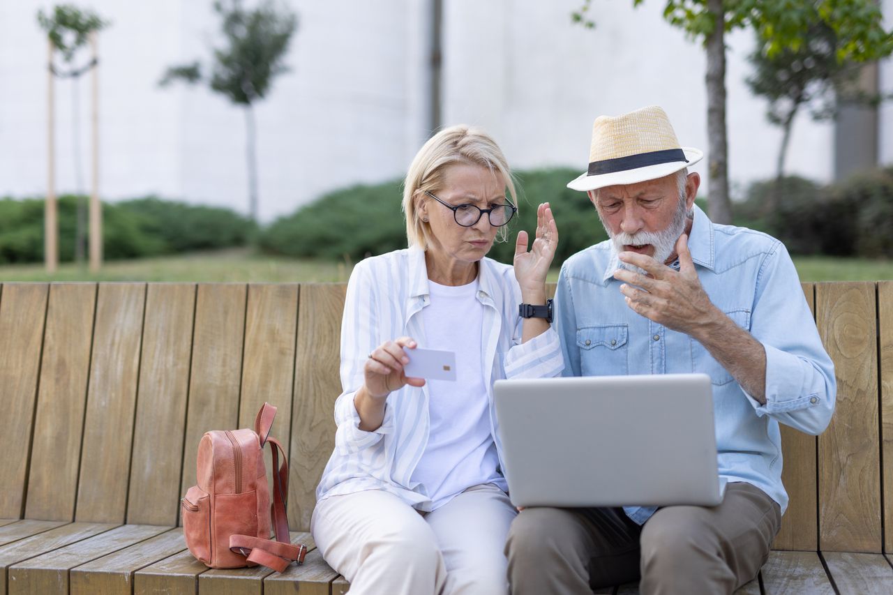 A mature couple sits closely on a wooden bench, showing concern as they look at a laptop, highlighting the reality of dealing with challenges together in an outdoor park setting.