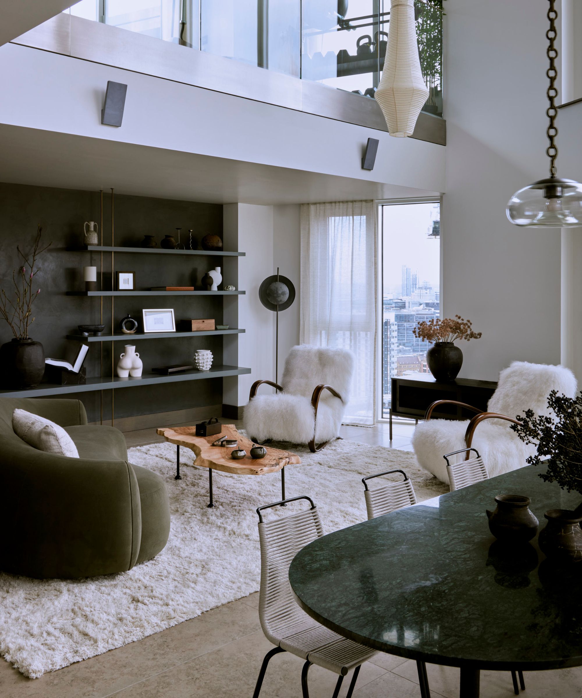 cozy modern open plan living room area with a dining table set up with a green marble table, modern chairs, a large living area with a white fluffy rug with a green velvet curved couch and sheepskin arm chairs