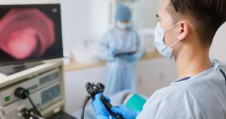 doctor wearing a surgical mask stands in front of a monitor while performing a colonoscopy on a patient that's out of frame. A nurse in scrubs stands in the background with a tablet