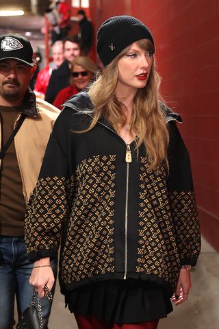 Taylor Swift has dark blonde hair, and is wearing a Louis Vuitton black and gold logo print jacket, a black beanie, and a Reputation necklace