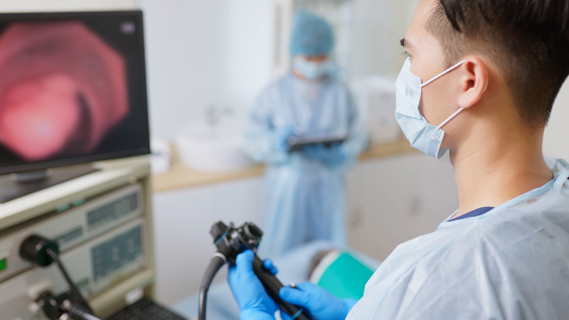 A person wearing scrubs and a face mask looks at a hospital monitor as a colonoscopy is performed.