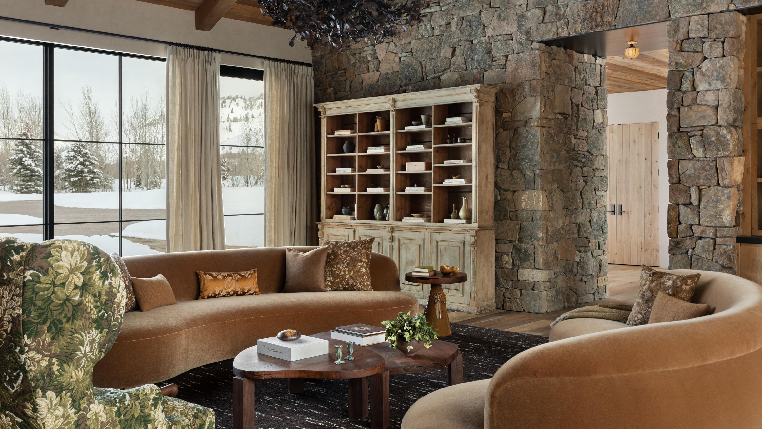 a stone clad living room with an open arched door, large glass doors looking out to a snowy yard, a rustic bookcase, with curved velvet couches