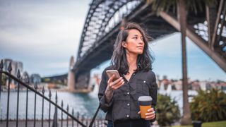 A young woman is using a phone in Sydney. The Sydney Harbour Bridge is behind her.