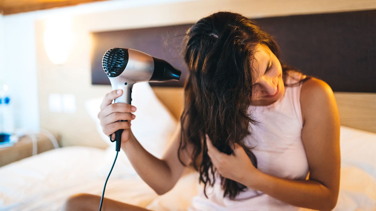 A woman who has just gotten out of the shower sits on her mattress and dries her hair