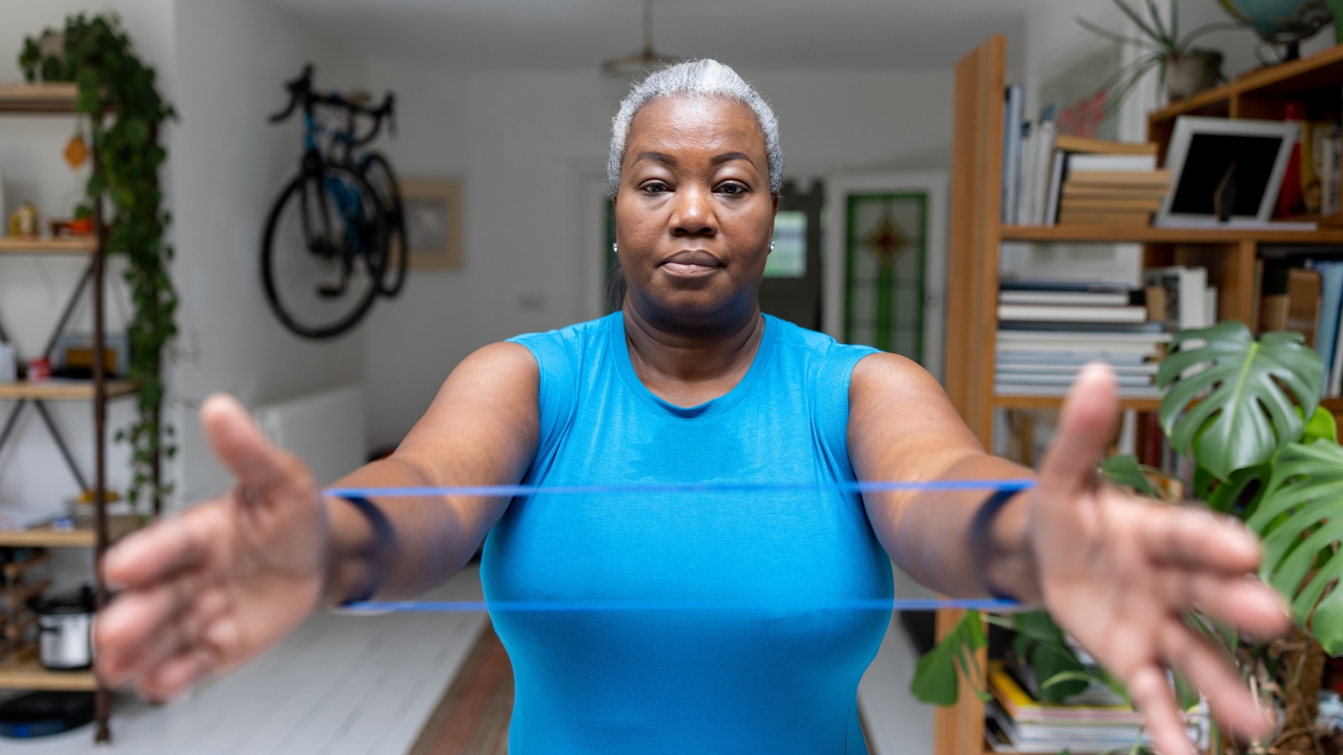 woman facing the camera wearing a bright blue sleeveless top holding her arms out toward the camera with a purple resistance band around her wrists. behind her is a home setting.