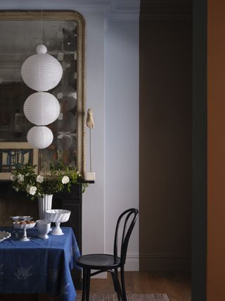 A dining room with the walls painted half white and half a dark mushroom brown. There is a dining table partially in view that has a royal blue tablecloth on it and one black dining chair at the head. There are multiple white but empty cake and food stands on the table and a white string of paper lanterns hanging like a pendant light.