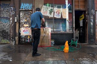 A man outside a building, using a hose to spray water on posters on the wall of a building