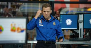 a man (taron egerton) wearing a tsa uniform listens to a phone call in the airport